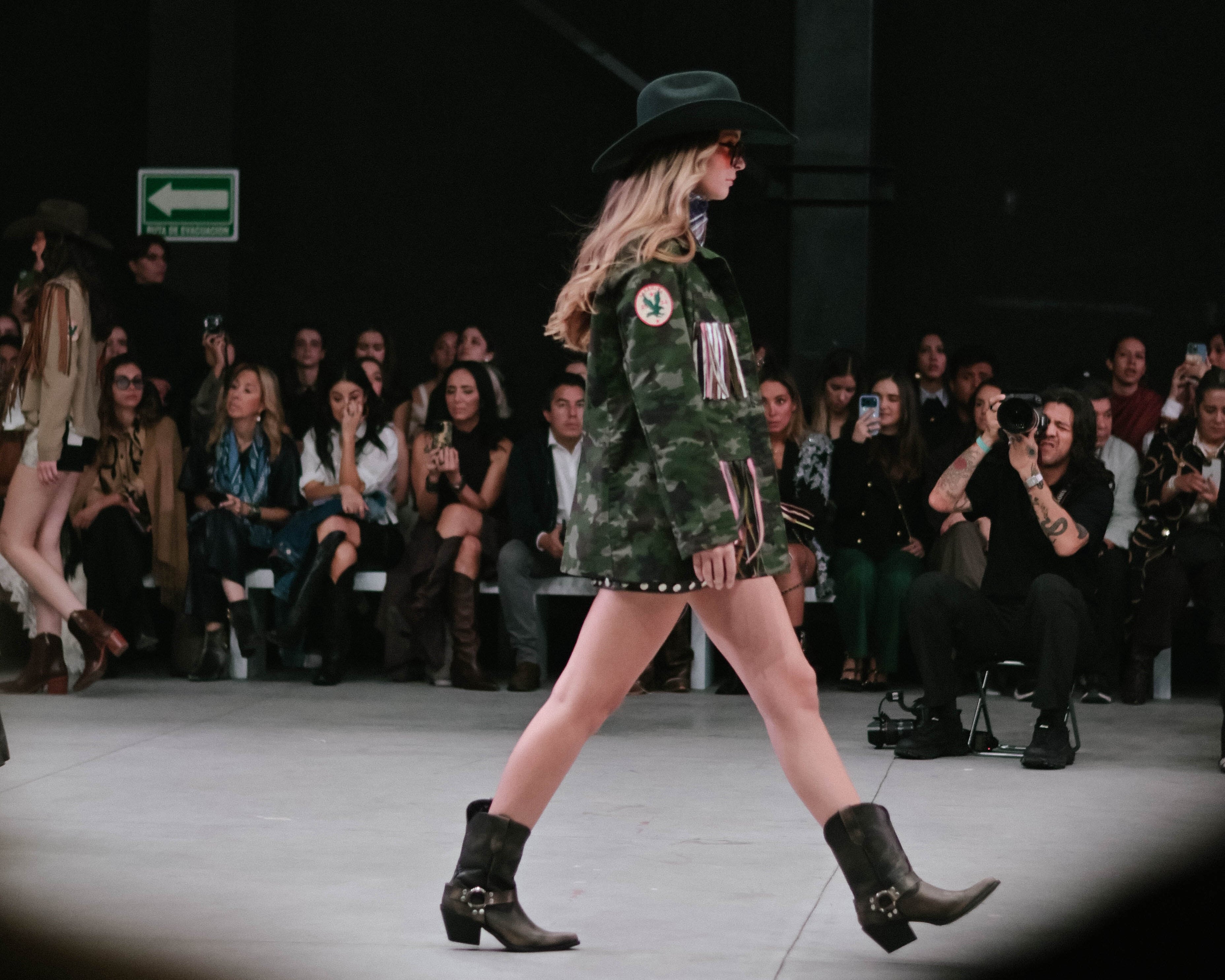 Model walking on a fashion show runway wearing a camouflage outfit and black boots.